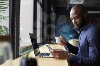 Mid adult black male creative sits by window having coffee, using a laptop and smartphone, side view