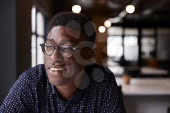 Young black male creative sitting at a desk in an office looking out of the window smiling, close up