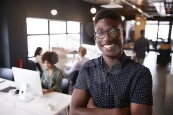 Millennial black male creative standing in a busy casual office, smiling to camera