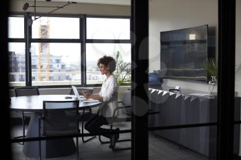 Millennial black businesswoman working alone in an office meeting room, seen through glass wall