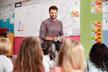 Male Teacher Reading Story To Group Of Elementary Pupils In School Classroom