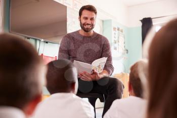 Male Teacher Reading Story To Group Of Elementary Pupils Wearing Uniform In School Classroom