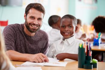Portrait Of Elementary School Teacher Giving Male Pupil Wearing Uniform One To One Support In Class