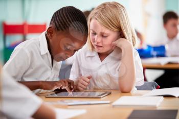 Two Elementary School Pupils Wearing Uniform Using Digital Tablet At Desk