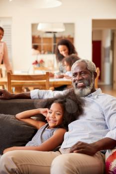 Grandfather With Granddaughter Sitting On Sofa At Home Watching Movie With Family In Background