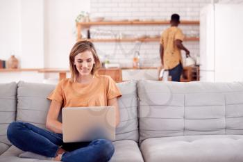Woman Relaxing Sitting On Sofa At Home Using Laptop Computer With Man In Kitchen Behind