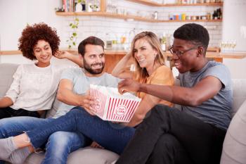 Group Of Friends Sitting On Sofa And Watching Movie At Home Whilst Eating Popcorn