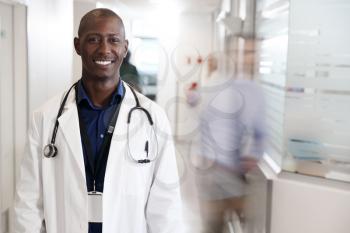 Portrait Of Smiling Male Doctor Wearing White Coat With Stethoscope In Busy Hospital Corridor