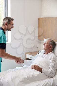 Surgeon Visiting And Shaking Hands With Senior Male Patient In Hospital Bed In Geriatric Unit