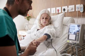 Male Nurse Taking Mature Female Patients Blood Pressure In Hospital Bed With Automated Machine