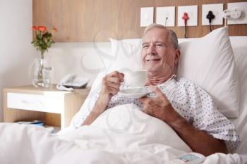 Male Senior Patient Lying In Hospital Bed Enjoying Hot Drink