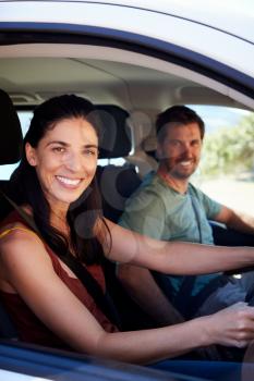 Mid adult white woman driving car, husband in front passenger seat smiling to camera, vertical
