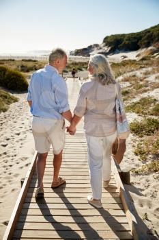 Senior white couple walking along wooden promenade on a beach holding hands, full length, back view