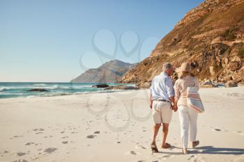 Senior white couple walking on a beach together holding hands, full length, back view