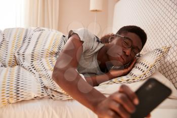 Millennial African American man wearing glasses, half asleep in bed holding smartphone, close up