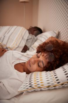 Millennial African American woman lying asleep in bed, her partner in the background, vertical