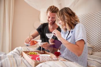 Millennial white couple celebrating with champagne,  breakfast and gifts in bed, close up