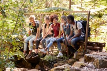 Five millennial friends sitting on a bridge in a forest talking during a hike, full length