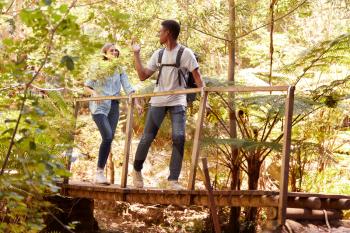 Two young adult friends hiking in a forest crossing a footbridge, full length