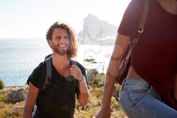 Young adult white man hiking with friends in countryside by the coast, smiling to camera, close up