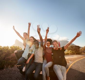 Five millennial friends on a road trip taking a break, leaning on the car waving to camera