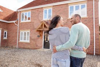 Rear View Of Couple Standing Outside New Home On Moving Day Looking At House