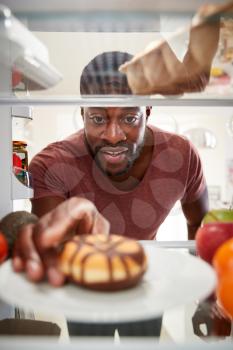 View Looking Out From Inside Of Refrigerator As Man Opens Door And Reaches For Unhealthy Donut