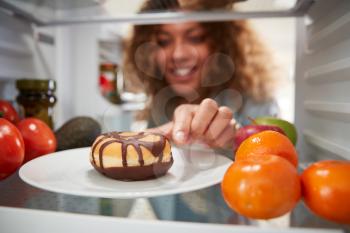 View Looking Out From Inside Of Refrigerator As Woman Opens Door And Reaches For Unhealthy Donut