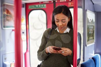 Female Passenger Standing By Doors In Train Looking At Mobile Phone