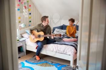 Single Father Playing Guitar With Son Who Drums On Cushion In Bedroom