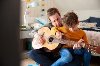 Single Father At Home With Son Teaching Him To Play Acoustic Guitar In Bedroom