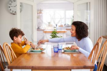 Single Mother Sitting At Table Eating Meal With Son In Kitchen At Home