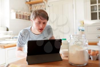 Young Man With Downs Syndrome Following Recipe On Digital Tablet To Bake Cake In Kitchen At Home