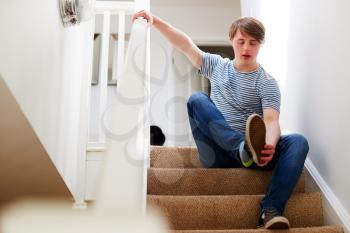 Young Downs Syndrome Man Sitting On Stairs Putting On Shoes At Home