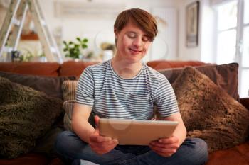 Young Downs Syndrome Man Sitting On Sofa Using Digital Tablet At Home