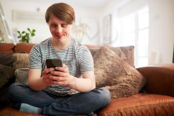 Young Downs Syndrome Man Sitting On Sofa Using Mobile Phone At Home