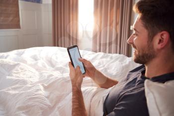 Man Sitting Up In Bed Looking At Mobile Phone After Having Woken Up