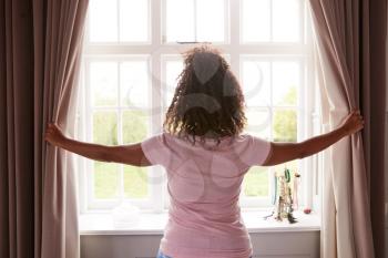 Rear View Of Woman Wearing Pajamas Opening Bedroom Curtains At Start Of New Day