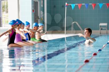 Male Coach In Water Gives Children Instructions In Swimming Lesson As They Sit On Edge Of Pool