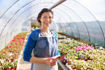 Portrait Of Mature Woman Working In Garden Center Greenhouse With Digital Tablet And Checking Plants