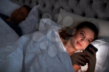 Woman Lying In Bed Checking Mobile Phone Whilst Man Sleeps Next To Her