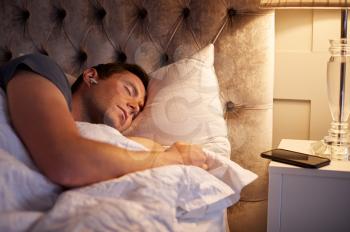 Man Sleeping In Bed Wearing Wireless Earphones Connected To Mobile Phone On Bedside Table