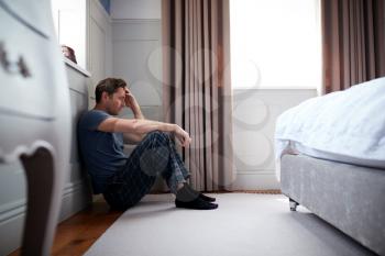 Depressed Man Wearing Pajamas Sitting On Floor Of Bedroom