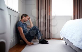 Depressed Man Wearing Pajamas Sitting On Floor Of Bedroom Holding Glass Of Whisky