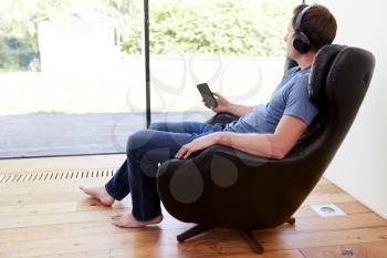 Man Relaxing In Chair At Home Streaming Music From Mobile Phone To Wireless Earphones