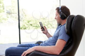 Man Relaxing In Chair At Home Streaming Music From Mobile Phone To Wireless Earphones