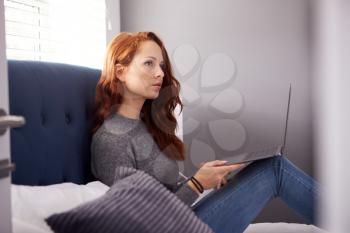 Female College Student In Shared House Bedroom Sitting On Bed With Laptop Studying