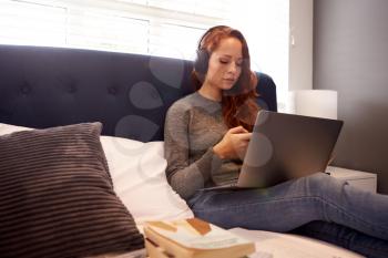 Female College Student Wearing Headphones Works On Bed In Shared House With Laptop And Mobile Phone