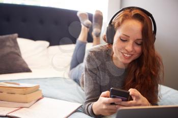 Female College Student Wearing Headphones Lies On Bed In Shared House With Mobile Phone Using Laptop