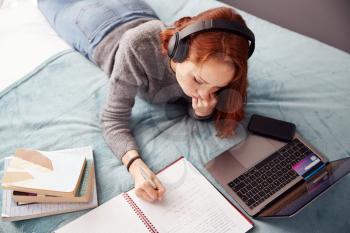 Looking Down On Female College Student Wearing Headphones Lying On Bed Working On Laptop